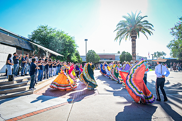 Students dance in colorful folklorico dresses, while mariachi musicians perform behind them