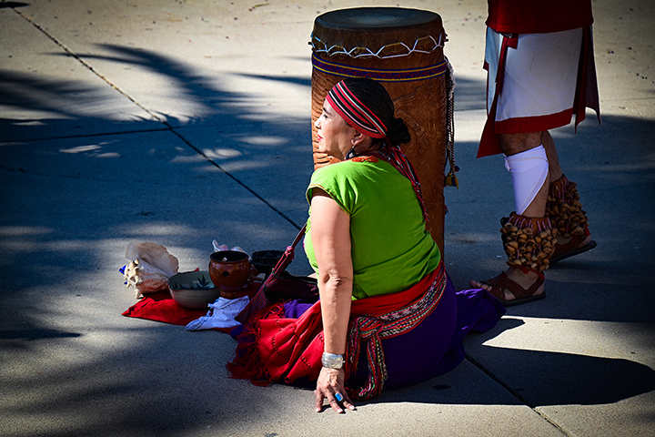 A woman in a green top and colorful headscarf sits on the ground next to a drum