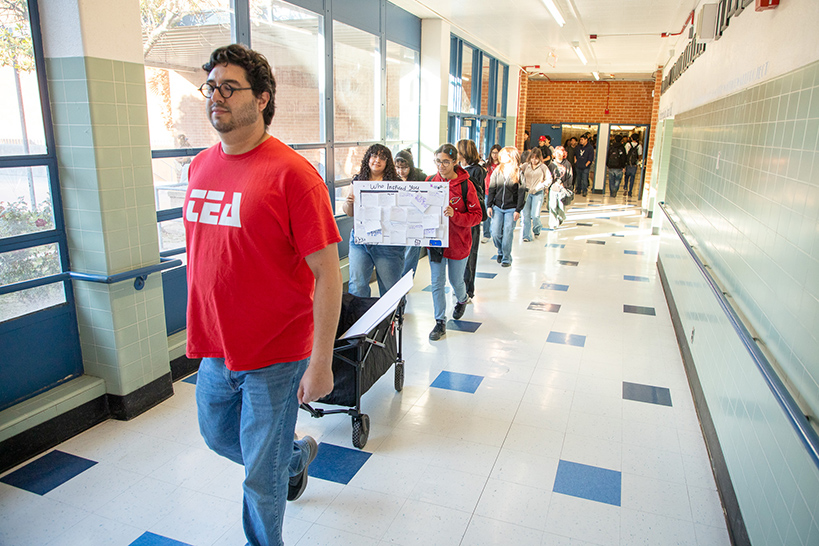 A man in a red shirt pulling a wagon with students holding signs as they walk behind them through the hallway