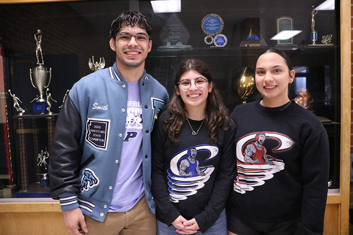 A teen boy and two teen girls smile in front of a trophy case