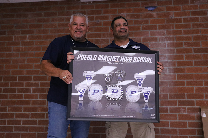 Two men smile as they hold up a framed posted of Pueblo High School's wrestling championship rings