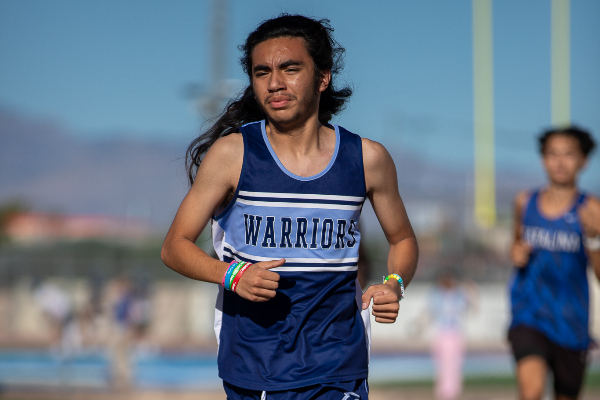 A teen boy with long dark hair wearing a blue Warriors singlet runs the cross country course